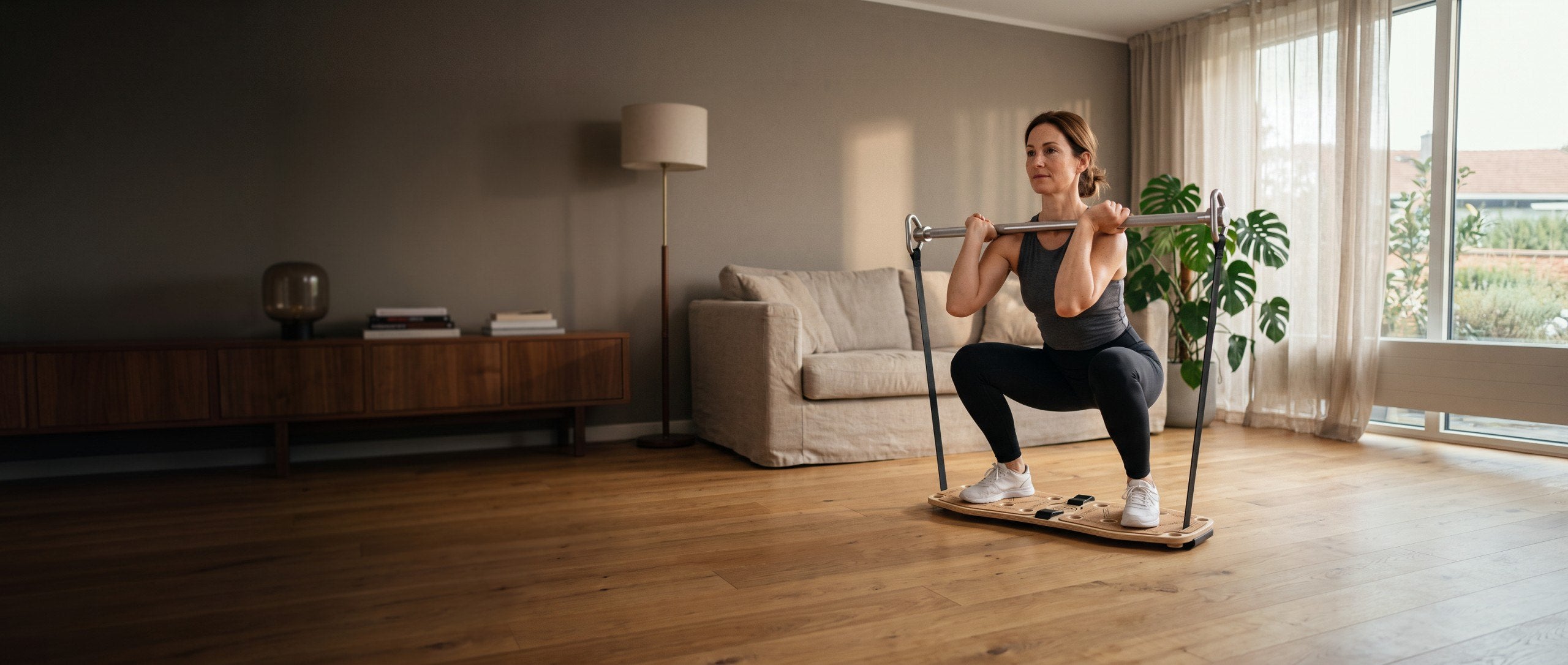 Frau macht im Wohnzimmer eine Kniebeuge auf dem Sportboard 4 Fitness, die Stange mit den Bändern vor den Schultern
